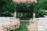 a gorgeous wedding ceremony space with an acrylic wedding arch covered with blush and pink blooms, with matching blush and pink blooms lining up the aisle