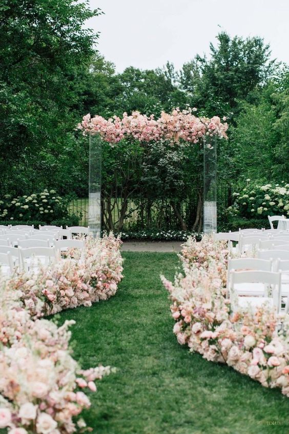 a gorgeous wedding ceremony space with an acrylic wedding arch covered with blush and pink blooms, with matching blush and pink blooms lining up the aisle
