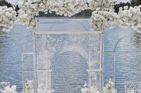 a jaw-dropping wedding ceremony space with a lake view, lush white floral lining up the aisle and covering the acrylic arch
