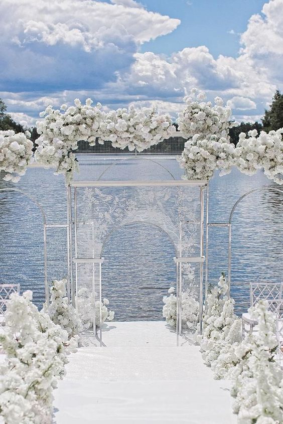 a jaw-dropping wedding ceremony space with a lake view, lush white floral lining up the aisle and covering the acrylic arch