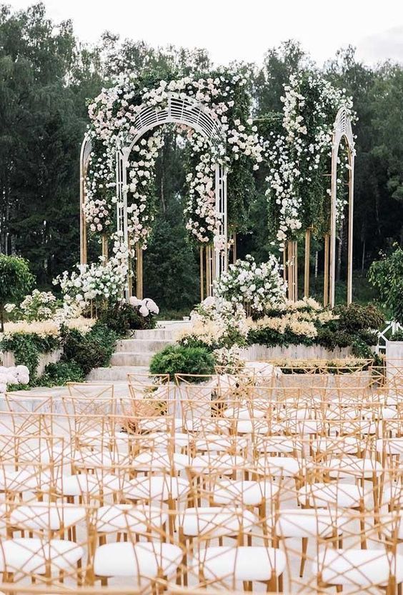 a jaw-dropping wedding ceremony space with a unique arbor with greenery and white and blush blooms and lots of grasses and greenery around it
