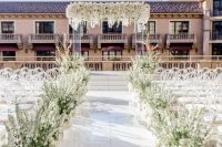 a lavish white wedding ceremony space with an acrylic wedding arch with white blooms, white floral arrangements and ghost chairs
