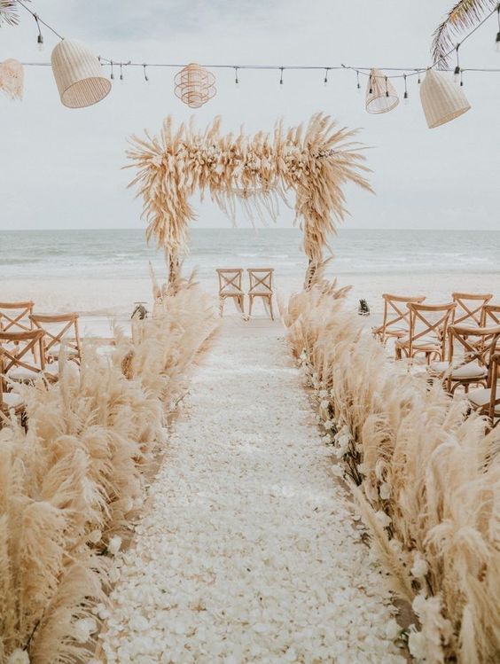 a luxurious boho wedding ceremony space on the beach, with pampas grass lining up the aisle and covering the arch