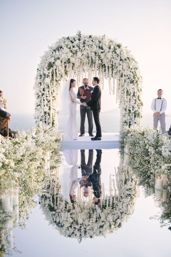 a luxurious wedding ceremony space with an incredibly lush white flower wedding arch and matching arrangements on the mirror floor
