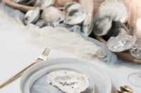 a neutral beach bridal shower table with watercolor plates, mussel shells, candles in glass candleholders and gold cutlery