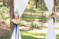 a outdoor rustic wedding arch decorated with white fabric and ribbons, with pastel and neutral blooms and greenery