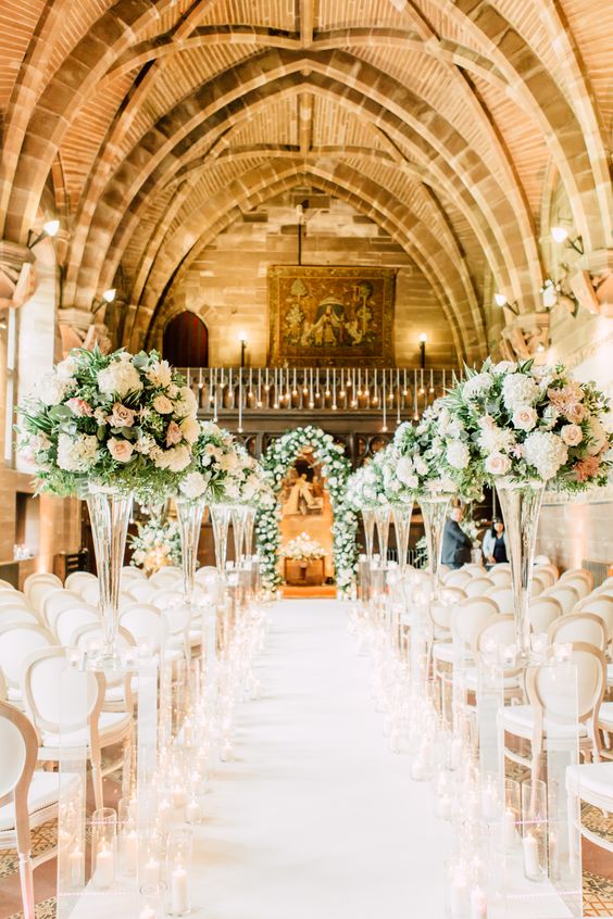 a refined wedding ceremony space with floral arrangements and greenery in tall clear vases, with a lush floral arch and candles