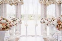 a refined wedding ceremony space with pillars and white blooms on top, floral arrangements in urns and a white runner