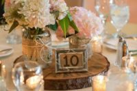 a rustic wedding centerpiece of a wood slice, a table number, pink and neutral blooms in jars wrapped with burlap