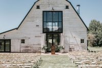 a serene outdoor barn wedding ceremony space with white benches and chairs, with white florals and curtains and no aisle decor