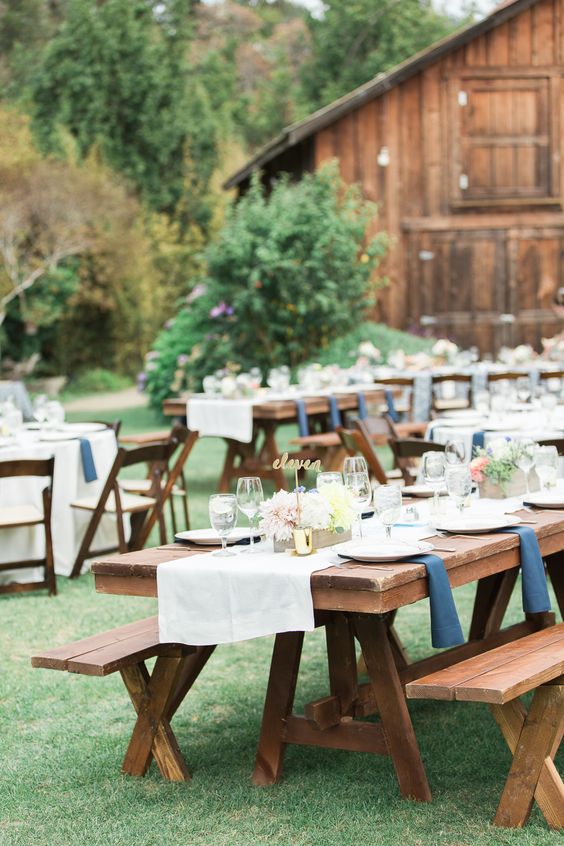 a stylish rustic outdoor barn reception space with stained tables and benches, with neutral and navy linens, with pastel blooms and gilded touches