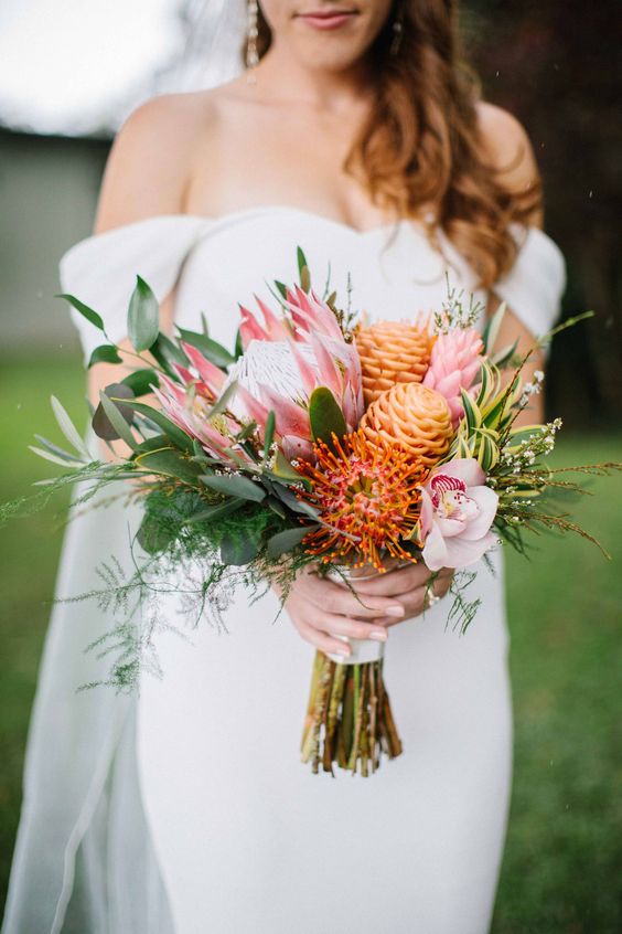 a stylish tropical wedding bouquet of king proteas, pincushion ones, some blush orchids, greenery and blooming branches