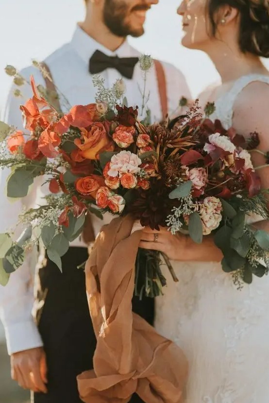 a super lush fall wedding bouquet of rust, blush, orange, burgundy blooms, lots of greenery and bright foliage, long tan ribbons is wow