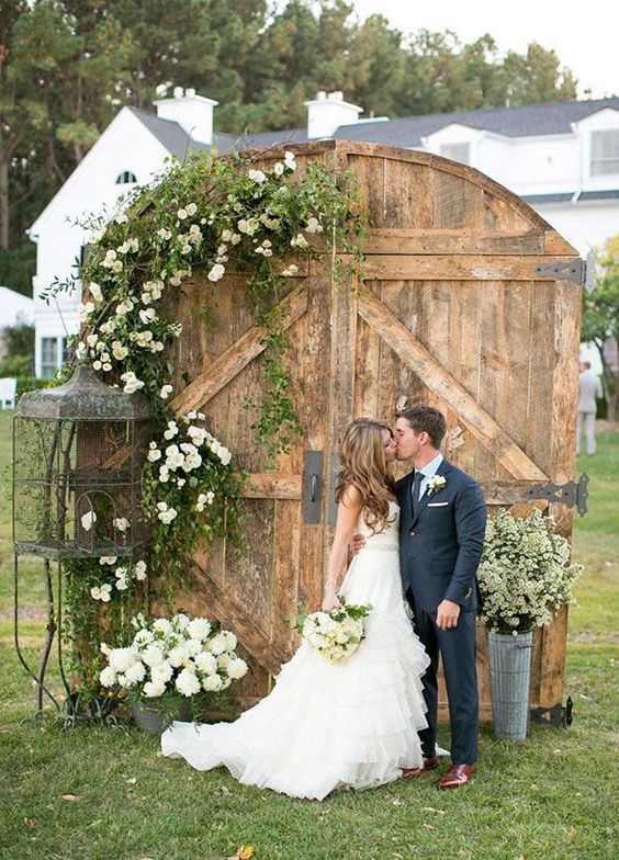 a vintage barn door wedding backdrop with greenery and white florals is a chic and beautiful idea for a barn wedding