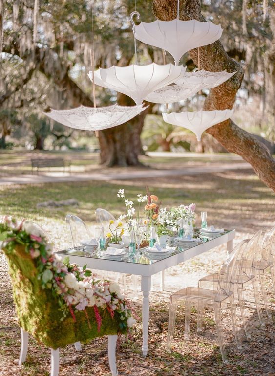 a whimsical garden bridal shower with lace umbrellas over the table, a glass table with wildflowers and a moss and bloom covered chair