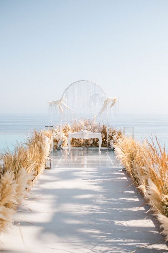 an airy wedding ceremony space with a clear backdrop with grass, pampas grass lining up the aisle and the backdrop