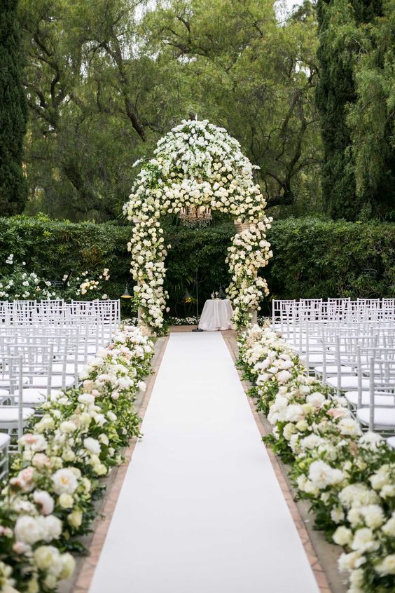 an elegant flower-filled wedding ceremony space with white and lush blooms lining up the aisle and a lush floral arch
