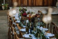 an outdoor barn wedding reception space with uncovered tables, neutral linens and a lush greenery runner plus blooms in a crate and string lights