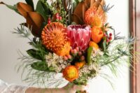 orange pincushion proteas, orange ranunculus, a king protea, magnolia leaves, greenery and waxflower compose a unique wedding bouquet