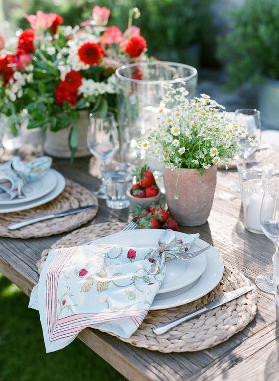 a beautiful summer wedding table setting with woven placemats, printed napkins, white and red blooms and some strawberry on the table