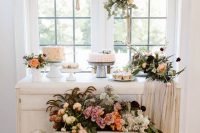 a beautiful white table with delicate neutral and pastel blooms and greenery and some cupcakes and cakes for a vintage bridal shower