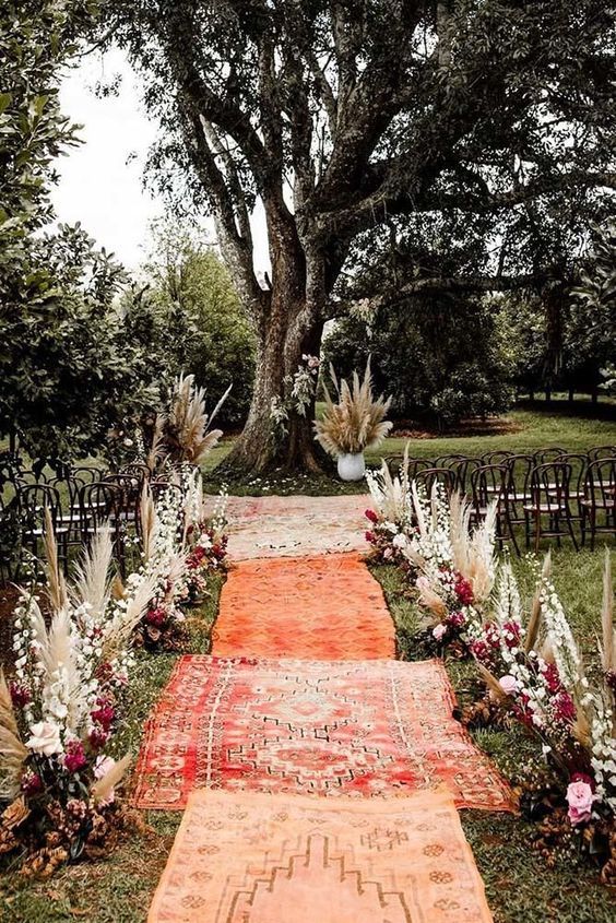 a brigth boho fall wedding aisle with greenery, pampas grass, fuchsia and blush blooms and white blooming branches plus rugs