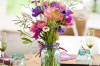a cluster wedding centerpiece of three mason jars, with bold blooms and greenery, purple ribbon and pebbles and candles in other jars