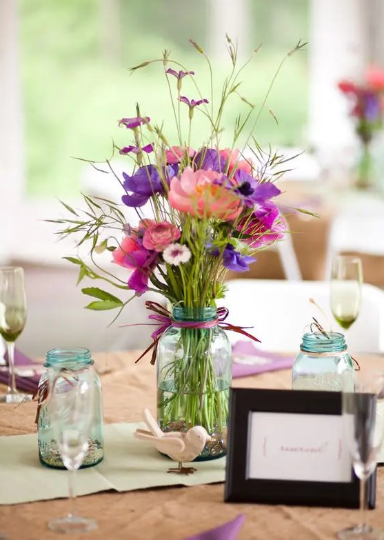 a cluster wedding centerpiece of three mason jars, with bold blooms and greenery, purple ribbon and pebbles and candles in other jars