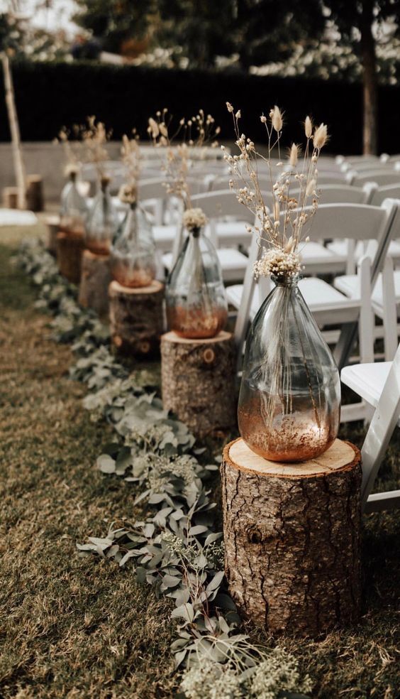 a cool fall wedding aisle lined up with greenery, with tree stumps, with vases with bunny tails and dried blooms is a chic idea