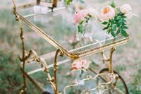 a gold and glass tiered cart with pink and bluhs blooms and greenery and candles can be used to serve desserts and drinks