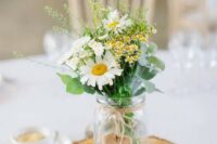 a lovely rustic wedding centerpiece of a tree slice, a mason jar with greenery and some wildflowers plus candles around