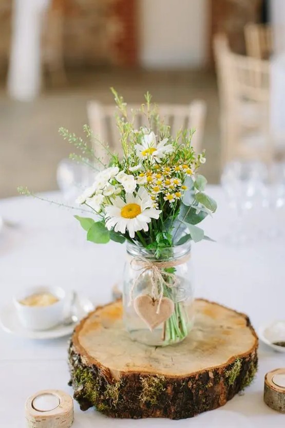 a lovely rustic wedding centerpiece of a tree slice, a mason jar with greenery and some wildflowers plus candles around