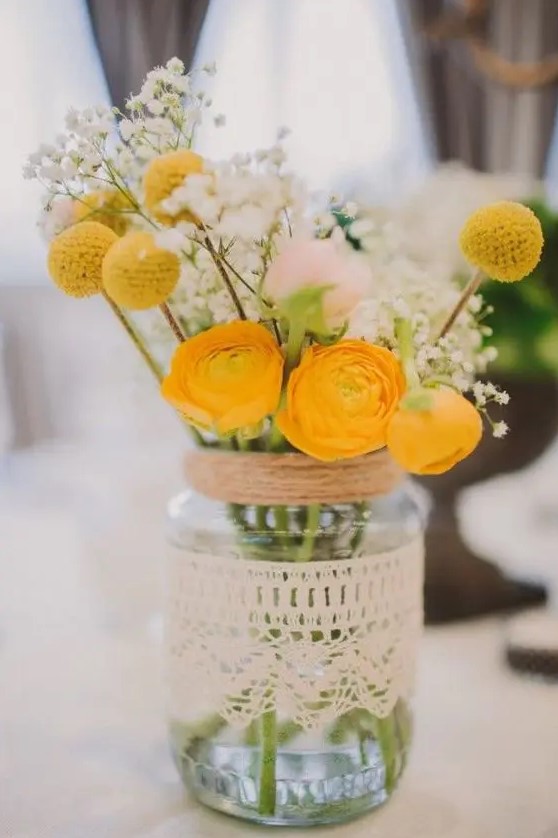a mason jar wrapped with a doily with a  floral arrangement with yellow ranunculus, baby's breath and billy balls