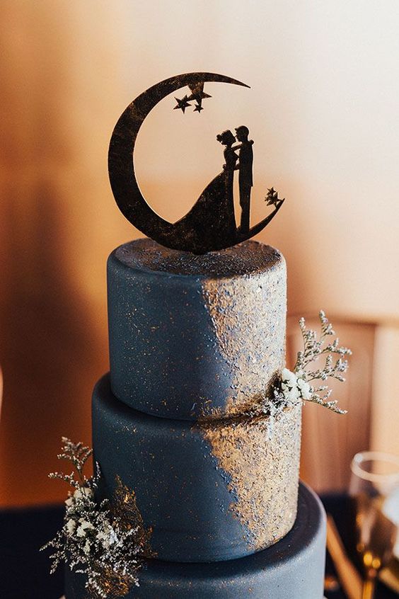 a navy wedding cake with gold touches, some fresh and dried blooms and a half moon silhouette and a couple is lovely
