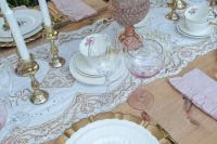 a pink vintage bridal shower tablescape with a lace runner, white candles, gold chargers and floral porcelain and gold cutlery