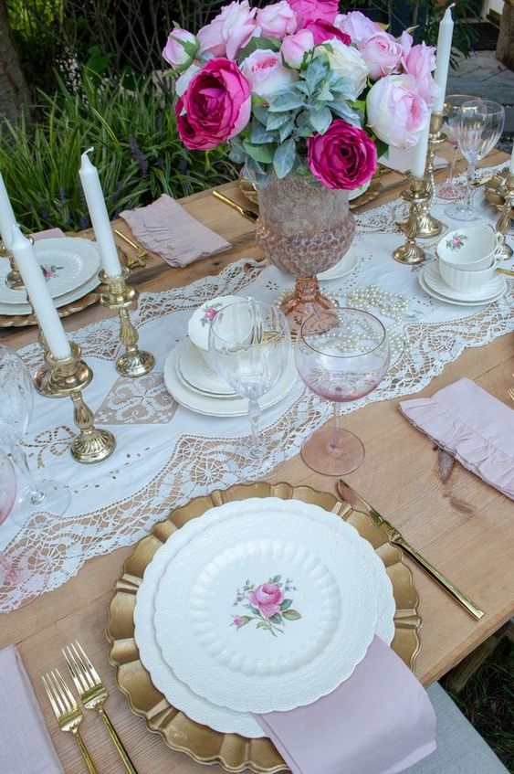 a pink vintage bridal shower tablescape with a lace runner, white candles, gold chargers and floral porcelain and gold cutlery