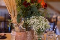a rustic wedding centerpiece of a tree slice, pumpkins, glittered jars, wheat, baby’s breath, orange and burgundy blooms, thistles and greenery