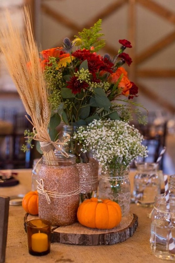 a rustic wedding centerpiece of a tree slice, pumpkins, glittered jars, wheat, baby's breath, orange and burgundy blooms, thistles and greenery