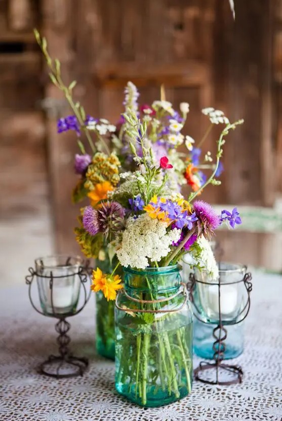 a summer wedding centerpiece of blue mason jars, with bold blooms and greenery, vintage candleholders with candles