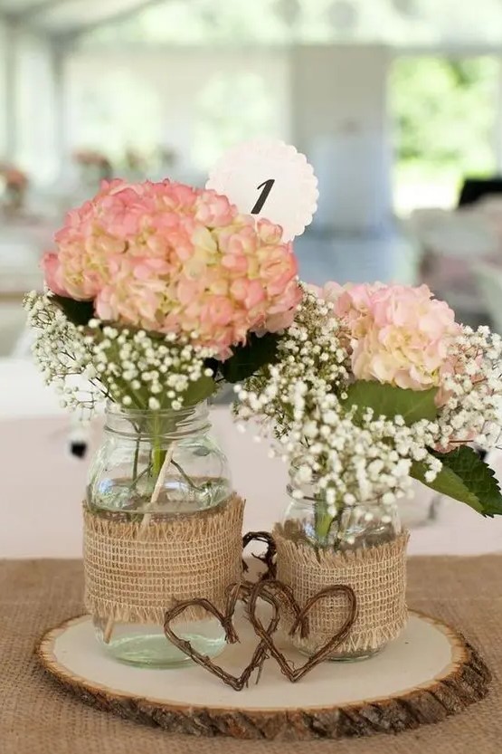 a tree slice with mason jars wrapped with burlap and with pink hydrangeas and white baby's breath, vine hearts for a rustic wedding