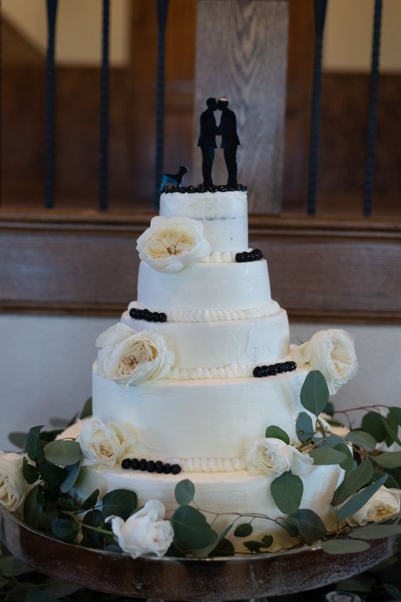 a white wedding cake topped with fresh berries and neutral blooms, with black silhouette cake toppers including the couple and their dog