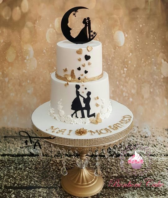 a white wedding cake with black and gold heart decor, with white sugar flowers, a black moon and couple silhouette cake topper