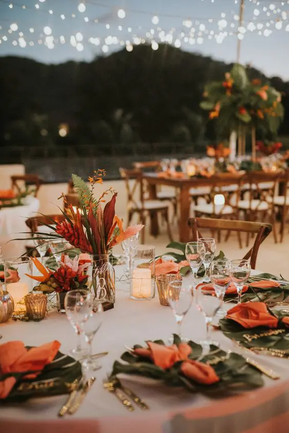a beautiful beach wedding table setting with fronds, orange blooms, candles and a neutral tablecloth is very elegant