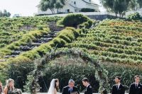 a beautiful fall vineyard wedding ceremony space with a trendy round greenery wedding arch and greenery decorating aisle chairs