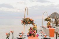 a bold and chic beach wedding reception space with neutral tablecloths and orange runners, orange blooms over the tables on them, colorful glasses
