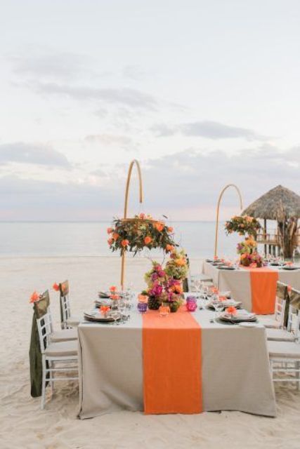 a bold and chic beach wedding reception space with neutral tablecloths and orange runners, orange blooms over the tables on them, colorful glasses