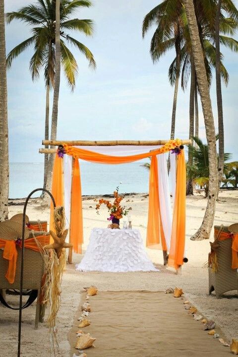 a bold tropical beach wedding ceremony space with an orange and white arch with blooms, orange blooms in a vase, wicker chairs with orange ribbon