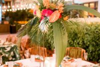 a bright tropical wedding reception space with woven placemats and printed napkins, pink glasses, a tall centerpiece with fronds, orange and pink bloom