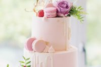 a light pink wedding cake with creamy drip, gold foil, bright blooms and greenery, pink macarons and a gold calligraphy topper