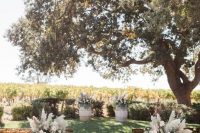 a lovely fall wedding ceremony space with a living tree, barrels with neutral blooms and pampas grass, simple chairs